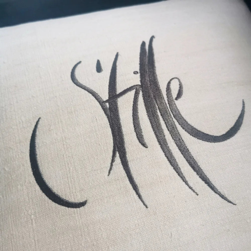 Detailaufnahme der anthrazitfarbenen Stickerei „Stille“ (Sign by Jwala®) auf naturcremer Bauernleinen-Kissenhülle mit sichtbarer Leinenstruktur