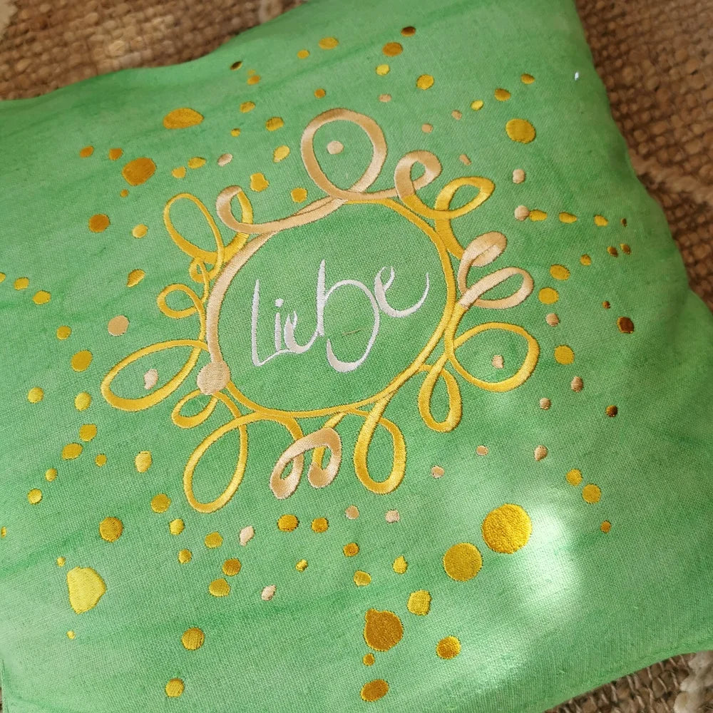 Wohnszene mit hellgrünem Bauernleinen-Sitzkissen „Liebe/Ornament“ als Blickfang auf einer Bank neben Decke und Kerze