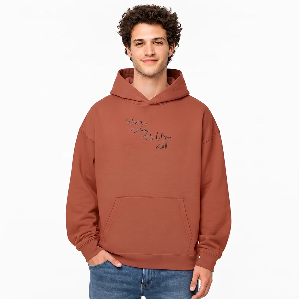 Junger Mann im terracottafarbenen Oversized-Hoodie mit einem mehrzeiligen, feinen Sign "Gehen, wohin das Leben will auf der Vorderseite vor weißem Hintergrund.