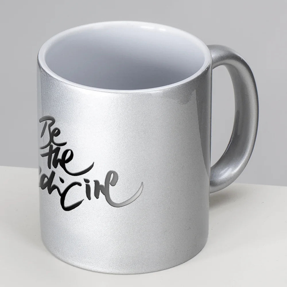 Silberne Metallic-Keramiktasse mit der schwarzen Aufschrift "Be the Medicine" in eleganter Schreibschrift Sign by Jwala® vor weißem Hintergrund.