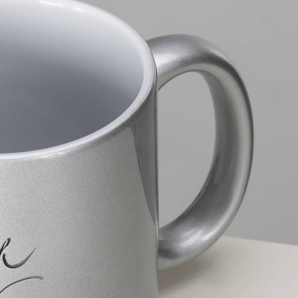 Makroaufnahme des ergonomischen Henkels und des oberen Randes einer silbernen Metallic-Kaffeetasse mit weißem Innenleben und Sign by Jwala®.