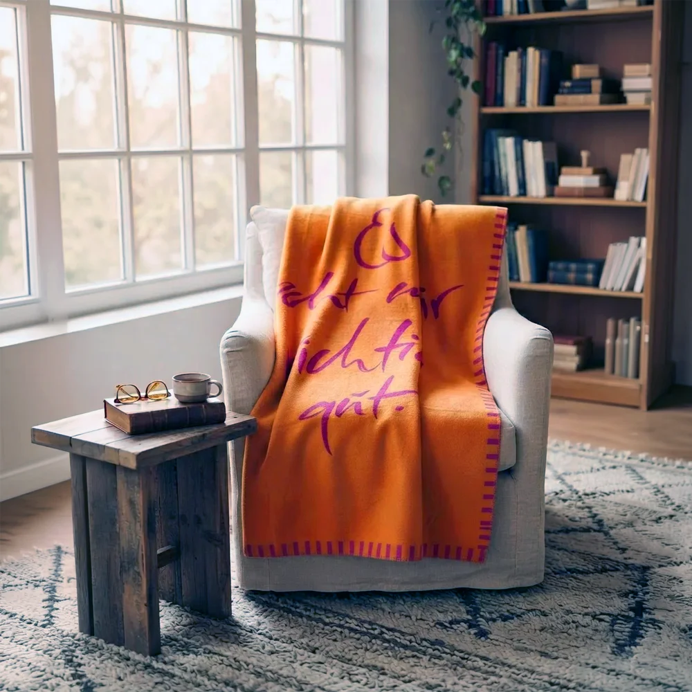 Stilvolle Wohnzimmer-Dekoration mit der Kuscheldecke „Es geht mir richtig gut“ auf einem Sessel neben einem Beistelltisch und Bücherregal.