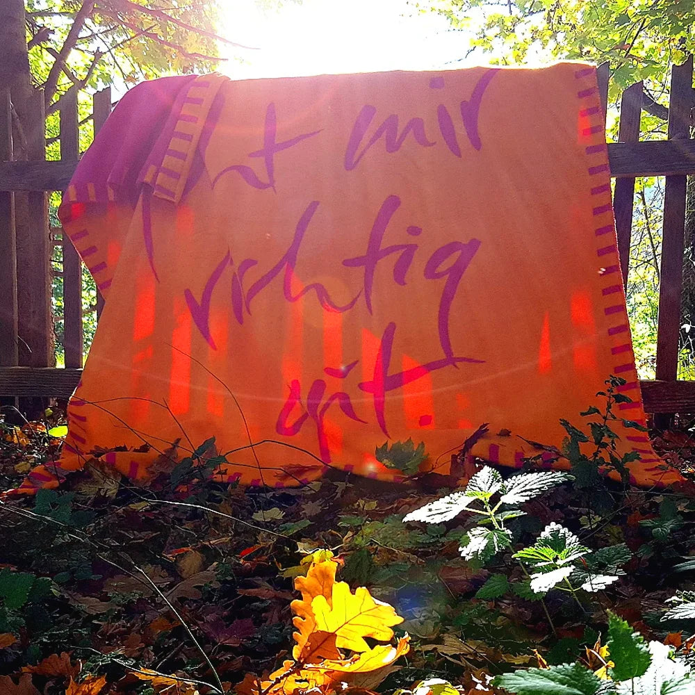 Orangefarbene Kuscheldecke von SignShopTirol mit dem pinken Schriftzug „Es geht mir richtig gut“ draußen an einem rustikalen Holzzaun im herbstlichen Garten.