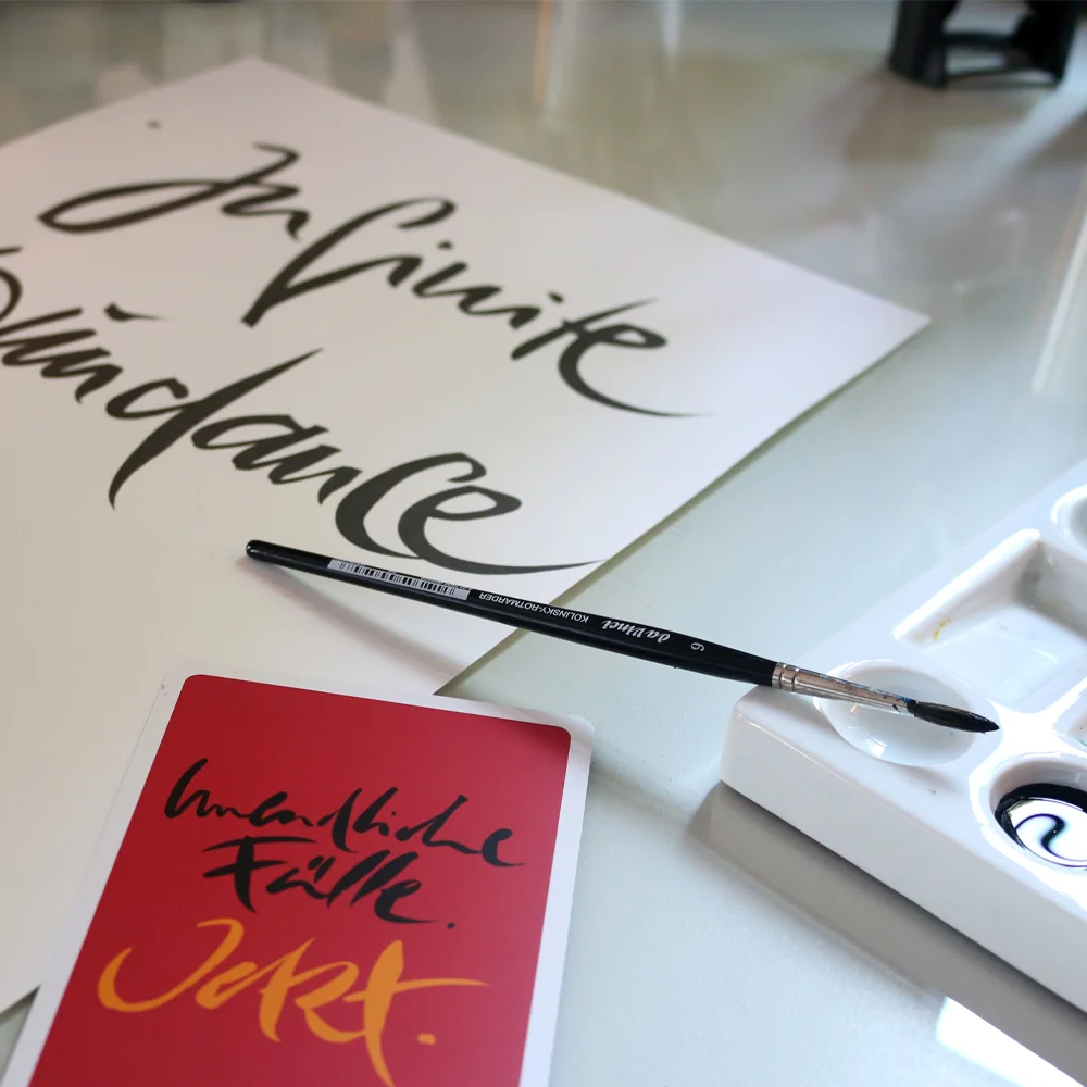 Handgemachte Kalligrafie auf Papier mit der Aufschrift „Infinite Abundance“ neben einem Pinsel, einer Farbpalette mit Tusche und einer roten SignCard mit dem deutschen Text „Unendliche Fülle. Jetzt.“