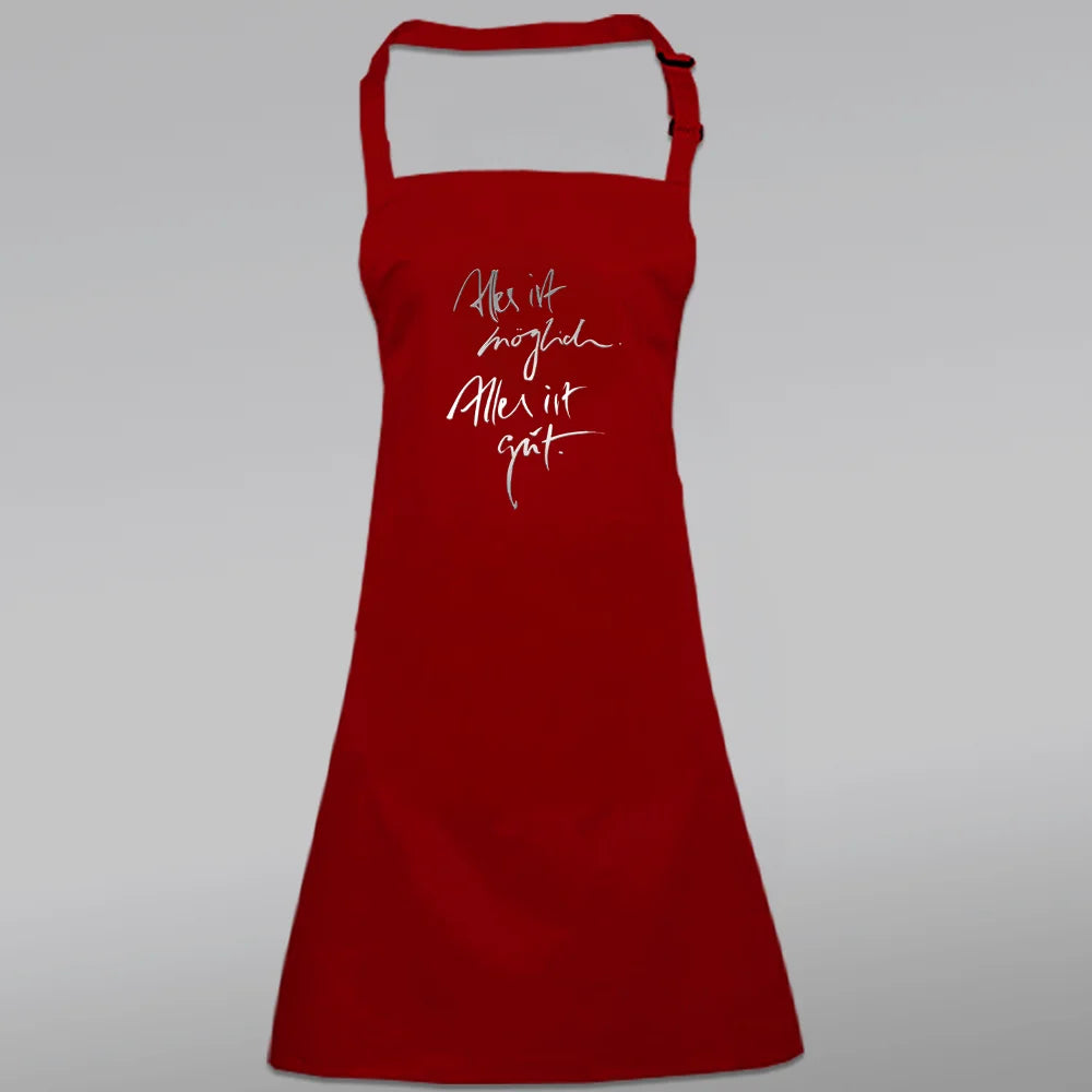 Rote Latzschürze bedruckt mit dem Jwala® Sign „Alles ist möglich. Alles ist gut.“ in silberner Schrift – exklusiv gestaltet von SignShop.tirol.