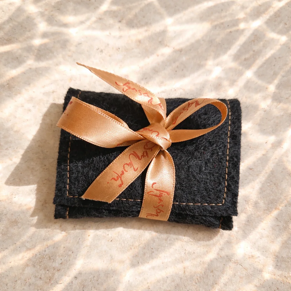 Ein kleines, dunkelgraues Etui aus 100 % Bauernleinen mit orangefarbenen Ziernähten, das mit einer edlen kupferfarbenen Satinschleife mit der Aufschrift „Signs by Jwala“ als Geschenk verpackt ist.