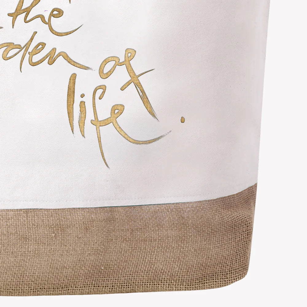 Nahaufnahme der Stoffstruktur einer weißen Strandtasche mit dem unteren Teil eines edlen, goldenen Aufdrucks, der die Worte „the garden of life.“ in eleganter Handschrift zeigt, direkt über dem naturfarbenen Juteboden.