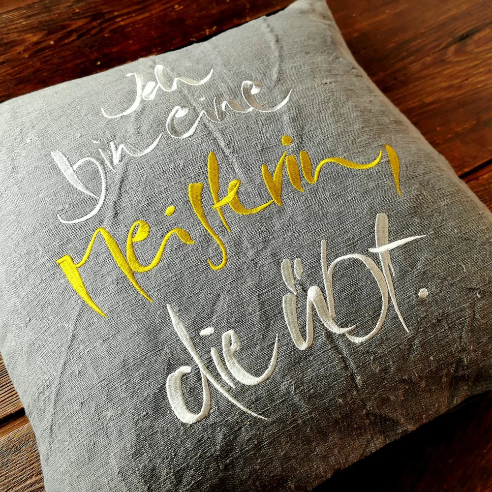 Flach liegende Kissenhülle aus Vintage-Bauernleinen mit silber-goldener Schrift „Ich bin eine Meisterin, die übt.“, sichtbare Leinenstruktur im Nahaufnahme-Fokus