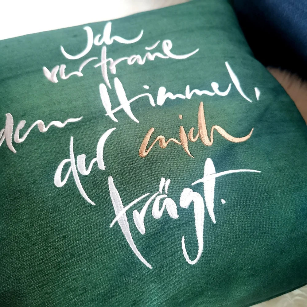 Detailaufnahme der mehrfarbigen Stickerei „Ich vertraue dem Himmel, der mich trägt“ (Sign by Jwala®) auf dunkelgrüner Bauernleinen-Kissenhülle