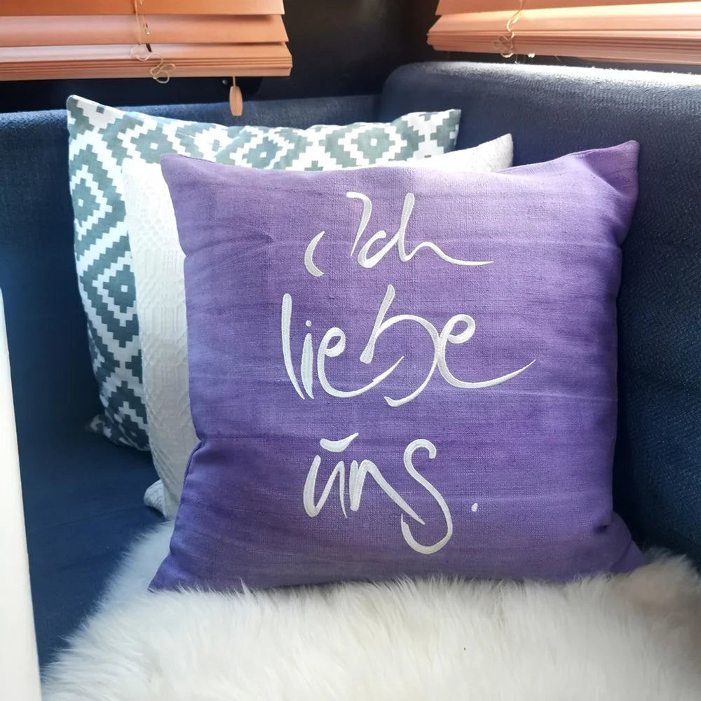Arrangierte hellviolette Bauernleinen-Kissenhülle „Ich liebe uns“ als liebevoller Akzent auf einem gemeinsamen Lieblingsplatz