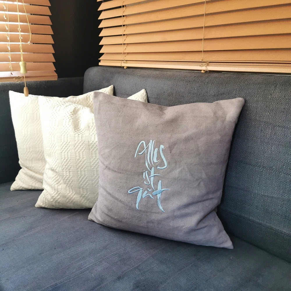 Graue Kissenhülle „Alles ist gut“ aus handgewebtem Vintage-Bauernleinen, 40x40 cm, mit hellblauer Stickerei auf einem Sofa dekoriert