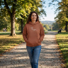 Lächelnde Frau geht im Park spazieren und trägt einen Heritage Brown Kapuzenpullover mit einem weißen, geschwungenen Sign "Alles ist möglich".