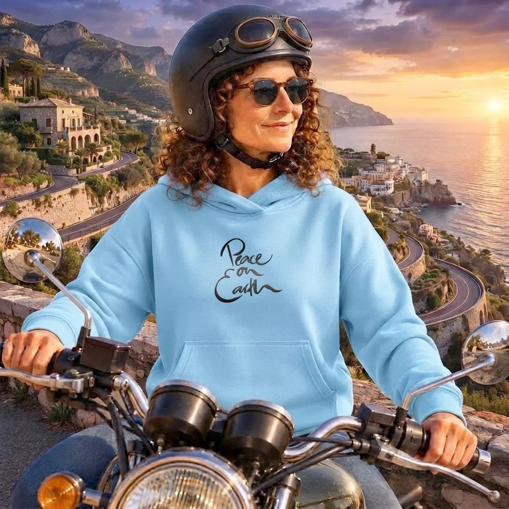 Frau mit lockigem Haar, Motorradhelm und Sonnenbrille blickt von einer Küstenstraße aufs Meer und trägt einen Kapuzenpullover in Blue Soul mit dem schwarzen Sign „Peace on Earth“ vom Signshop Tirol.