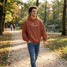 Lächelnder Mann geht im Park spazieren und trägt einen kastanienbraunen Oversized-Hoodie mit einem weißen, geschwungenen Sign "Alles ist Möglich".