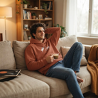 Entspannter Mann sitzt mit einer Kaffeetasse auf dem Sofa und trägt einen terracottafarbenen Kapuzenpullover mit weißem Sign #6oo vom Signshop Tirol.