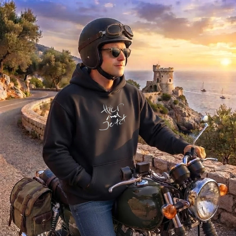Mann mit Motorradhelm und Sonnenbrille sitzt auf einem Classic-Bike an einer Küstenstraße und trägt einen schwarzen Kapuzenpullover mit dem weißen Schriftzug-Sign „Hier und Jetzt“