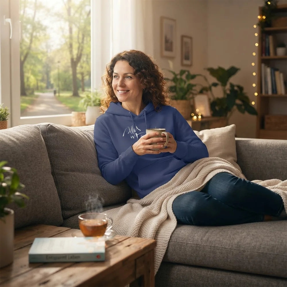 Entspannte Frau sitzt mit einer Tasse Tee auf dem Sofa und trägt einen blauen Damen-Hoodie mit weißem Schriftzug-Sign.
