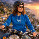 Lächelnde Frau auf einem Motorrad vor malerischer Küstenkulisse im dunkelblauen Hoodie mit dem weißen, modernen Sign „Jetzt“.