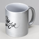 Silberne Metallic-Keramiktasse mit der schwarzen Aufschrift "Be the Medicine" in eleganter Schreibschrift Sign by Jwala® vor weißem Hintergrund.