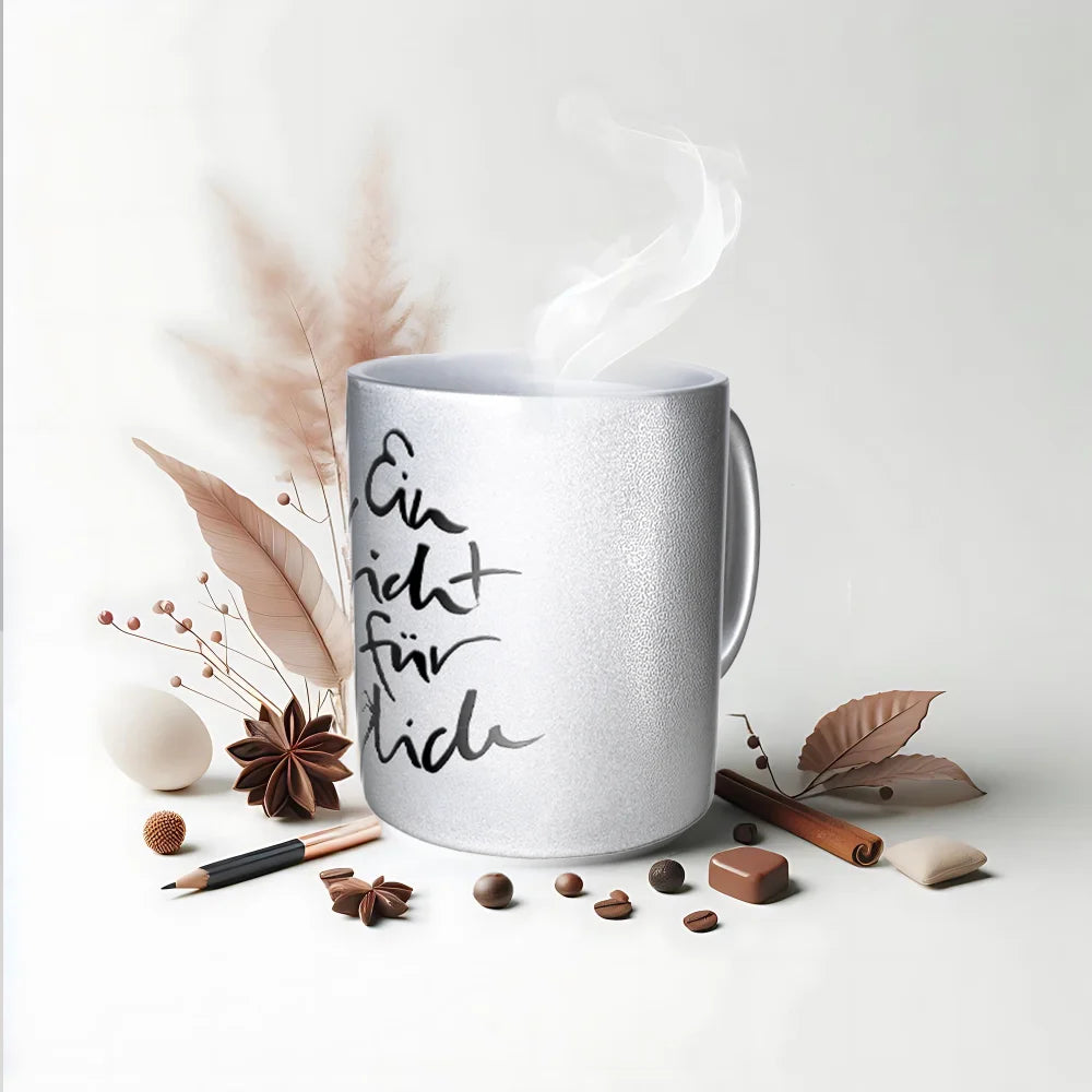 Silber glänzende Keramiktasse mit dem vertikalen schwarzen Schriftzug Sign by Jwala® „Ein Licht für dich“ in geschwungener Handschrift. Aus der Tasse steigt weißer Dampf auf. Sie steht auf einem weißen Untergrund, umgeben von herbstlichen Dekorationen wie Zimtstangen, Sternanis, Kaffeebohnen und getrockneten Gräsern.