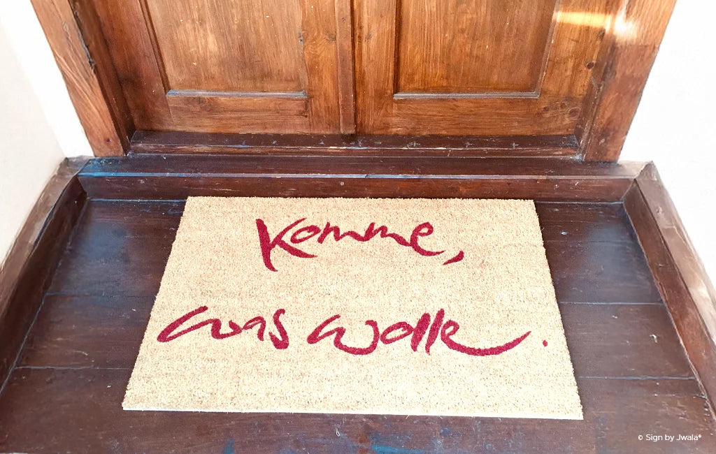 Das Bild zeigt einen Türteppich mit der aufgestickten Botschaft „Komme, was wolle“. Der Teppich liegt einladend vor einer Tür und vermittelt Zuversicht, Herzlichkeit und einen liebevollen Willkommensgruß.