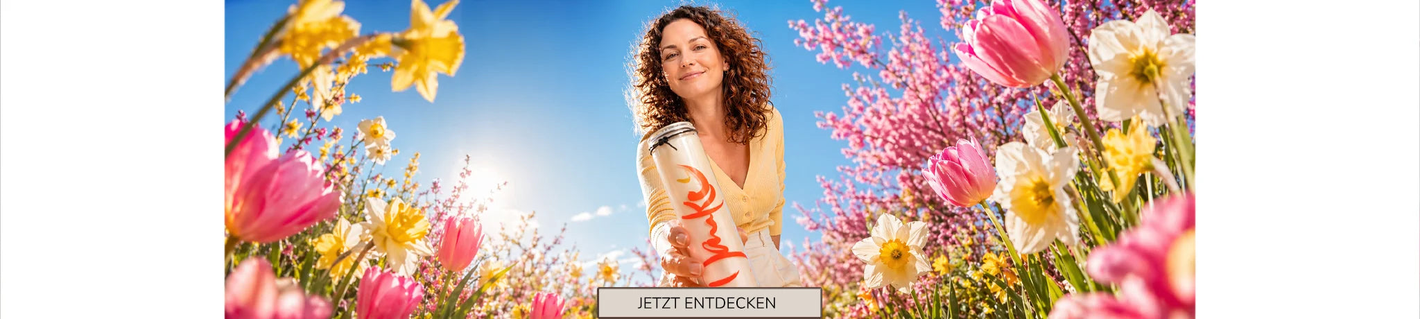 Frühlingshafter Shop-Header: Eine lächelnde Frau inmitten eines bunten Blumenmeers aus Tulpen und Narzissen hält ein SIGN-Lichtglas mit der Aufschrift „Danke“ in die Kamera. Mittig unten befindet sich ein Button mit der Aufschrift „JETZT ENTDECKEN“.