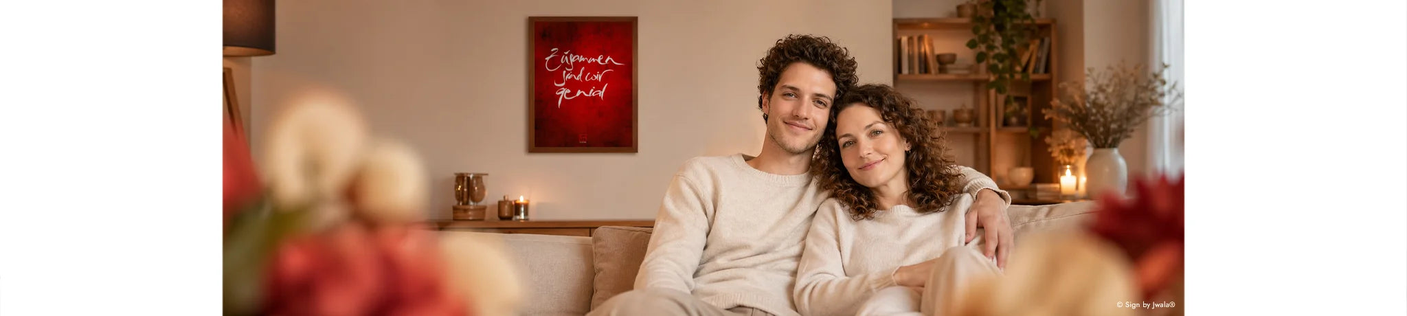 „Lifestyle-Aufnahme eines Paares in heller Kleidung, das entspannt auf einem beigen Sofa sitzt. Im Hintergrund hängt ein gerahmtes, dunkelrotes Poster aus dem SignShop Tirol mit dem kalligrafischen Schriftzug ‚Zusammen sind wir genial‘. Die Szene ist warm beleuchtet und mit natürlichen Holzregalen sowie Pflanzen dekoriert.“
