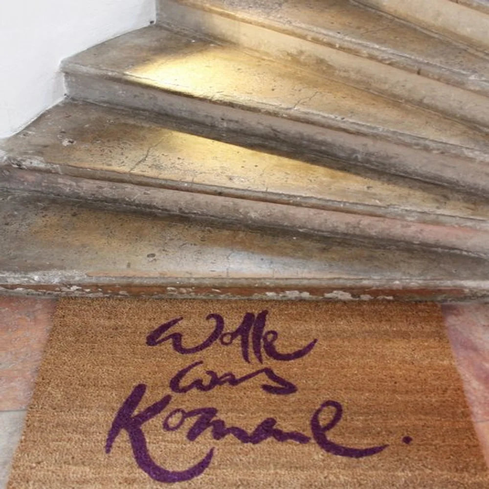 Handgefertigte Fußmatte aus 100 % Kokos mit inspirierendem Sign „Wolle, was komme“. In Violett, für Innenbereiche und überdachte Eingänge.