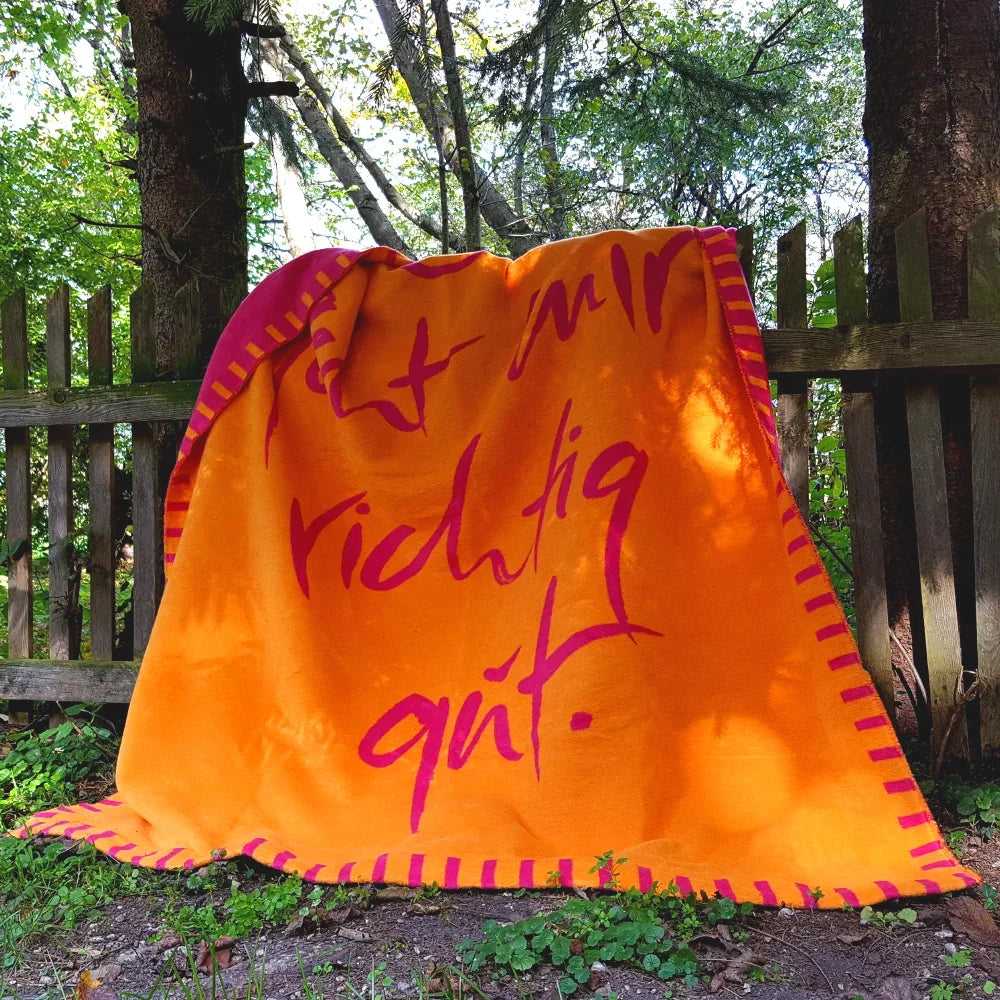 Kuscheldecke aus einer Vorarlberger Manufaktur in Orange und Fuchsia, angelehnt an einen Holzzaun im herbstlichen Garten.