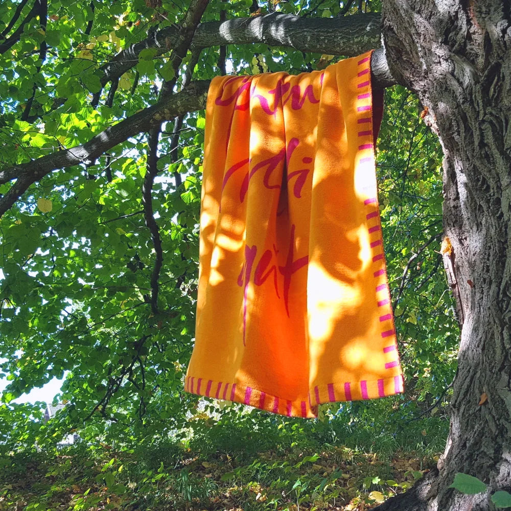 Die farbenfrohe Wohndecke in Orange und Fuchsia hängt dekorativ über einem Ast eines grünen Baumes in der Natur.