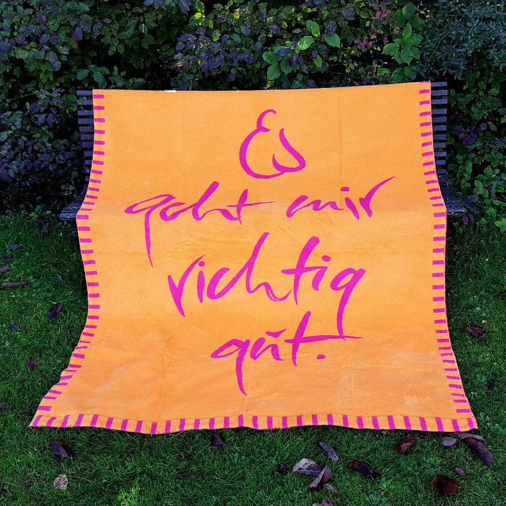 Kuscheldecke „Es geht mir richtig gut“ aus Baumwolle flach ausgebreitet auf einer Gartenbank im grünen Gras.