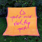 Kuscheldecke „Es geht mir richtig gut“ aus Baumwolle flach ausgebreitet auf einer Gartenbank im grünen Gras.