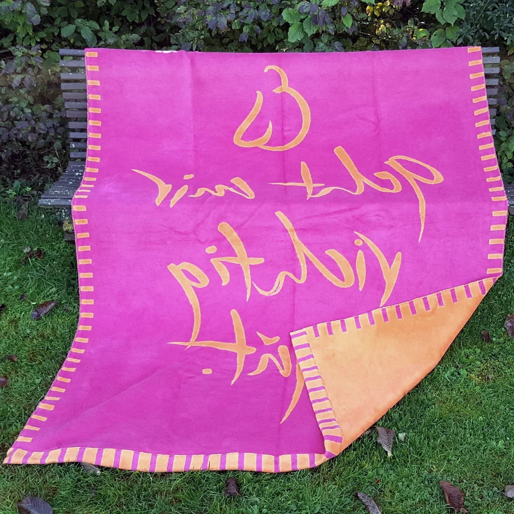 Spiegelverkehrte Rückseite der Decke in Fuchsia-Pink mit orangefarbener Jwala-Schrift auf einer Wiese im Freien.