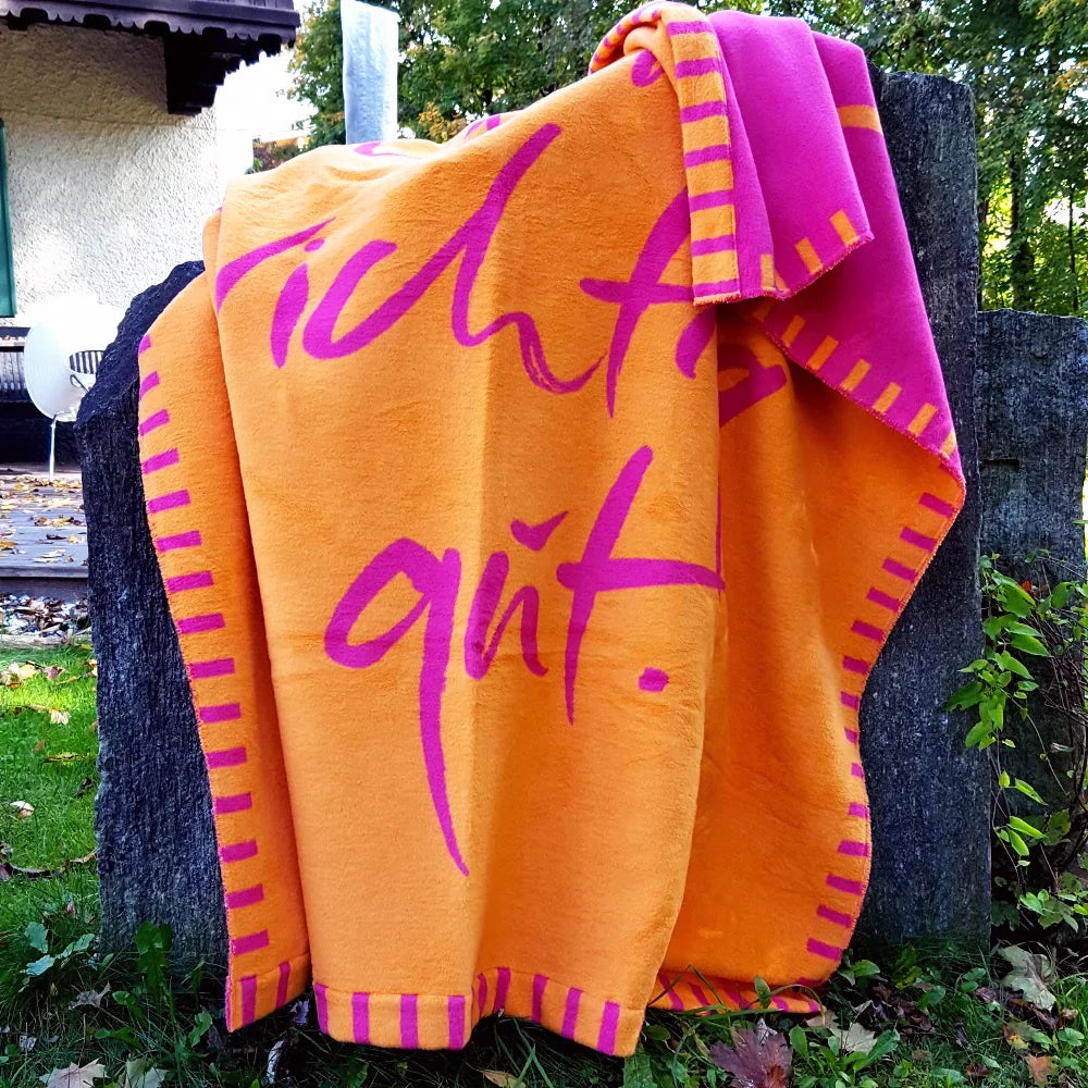 Kuscheldecke aus einer Vorarlberger Manufaktur in Orange und Fuchsia, angelehnt an einen Holzzaun im herbstlichen Garten.