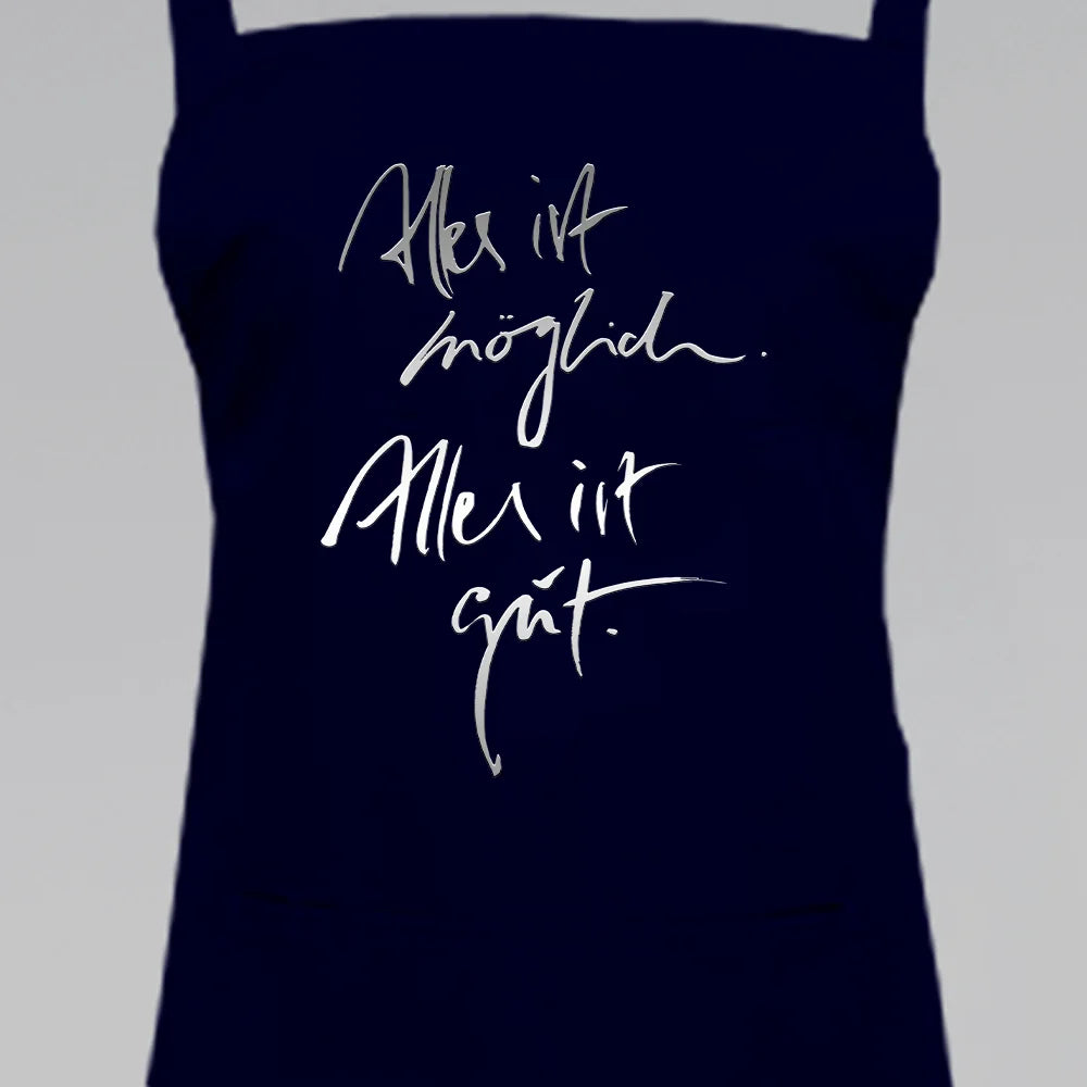 Schwarze Latzschürze mit dem silbernen Jwala® Sign „Alles ist möglich. Alles ist gut.“ in feiner Pinselschrift – hochwertige Kochschürze von SignShop.tirol.