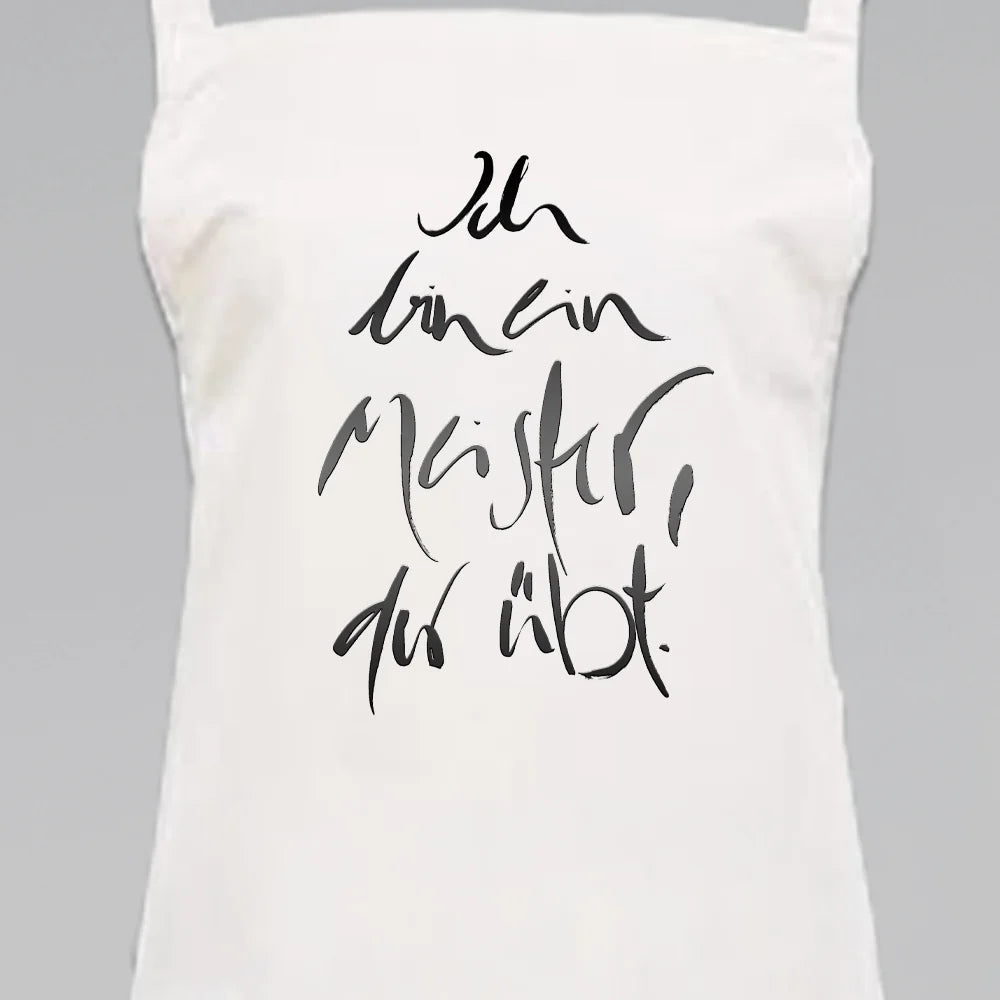 Weiße Latzschürze mit dem Titan farbenem Jwala® Sign „Ich bin ein Meister, der übt“ in ausdrucksstarker Kalligrafie – personalisierte Schürze online kaufen bei SignShop.tirol.