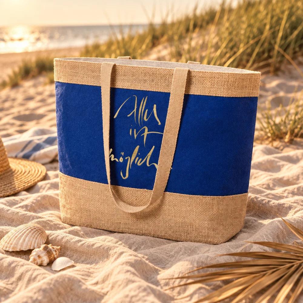 Eine zweifarbige Strandtasche aus naturfarbener Jute und blauem Canvas steht im weichen Sand an einem Dünenstrand bei Sonnenuntergang. Auf dem blauen Stoff prangt der goldene Schriftzug „Alles ist möglich“. Im Vordergrund liegen dekorative Muscheln auf einem Strandtuch.