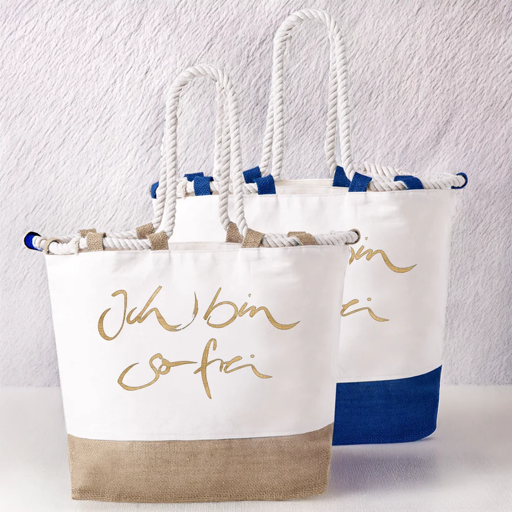 Zwei weiße Strandtaschen aus Baumwolle mit dicken, hellen Kordelgriffen. Die vordere Tasche hat einen naturfarbenen Juteboden und den goldenen Schriftzug „Ich bin so frei“, die hintere einen blauen Juteboden mit der Aufschrift „Alles ist möglich“