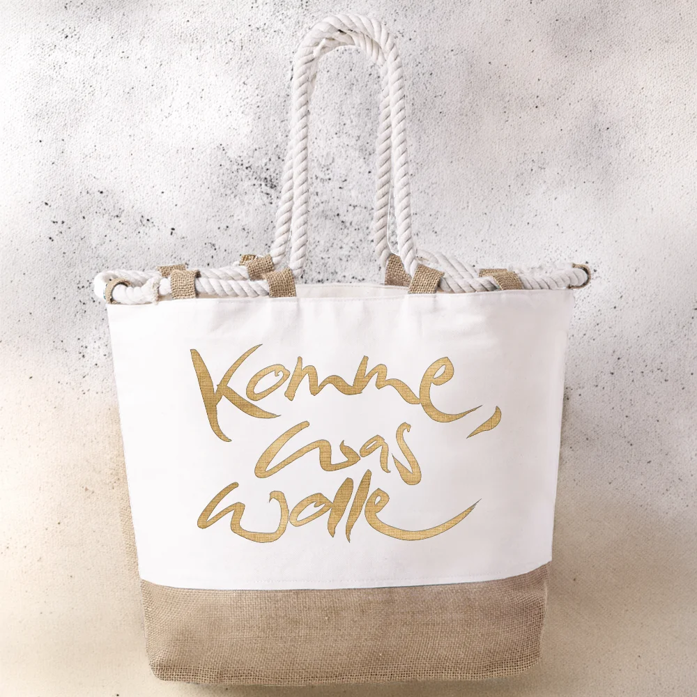 Weiße Strandtasche aus Baumwolle und naturfarbenem Juteboden mit dicken, hellen Kordelgriffen. Auf der Vorderseite ist der Spruch „Komme, was wolle“ in glänzender, goldener Schreibschrift aufgedruckt.
