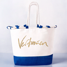 Veredelte Strandtasche aus weißer Baumwolle mit einem kontrastierenden, tiefblauen Juteboden und weißen Kordelgriffen. Die Vorderseite ist mit dem goldenen Wort „Vertrauen“ bedruckt.
