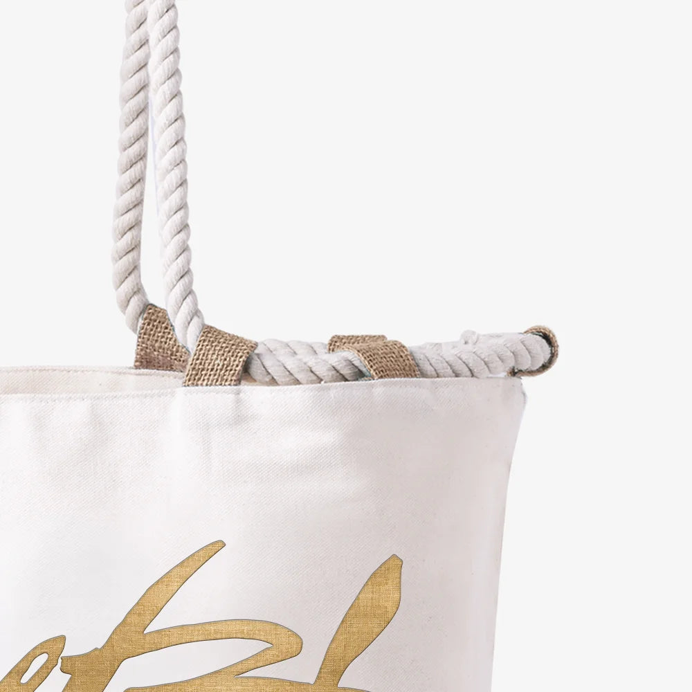 Detailaufnahme einer weißen Canvas-Strandtasche. Zu sehen ist der obere Rand mit den dicken, geflochtenen Kordelgriffen, die durch robuste Schlaufen aus naturfarbener Jute geführt sind, sowie ein Teil eines goldenen Schriftzugs.