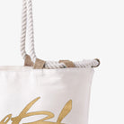 Detailaufnahme einer weißen Canvas-Strandtasche. Zu sehen ist der obere Rand mit den dicken, geflochtenen Kordelgriffen, die durch robuste Schlaufen aus naturfarbener Jute geführt sind, sowie ein Teil eines goldenen Schriftzugs.