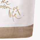 Nahaufnahme der Stoffstruktur einer weißen Strandtasche mit dem unteren Teil eines edlen, goldenen Aufdrucks, der die Worte „the garden of life.“ in eleganter Handschrift zeigt, direkt über dem naturfarbenen Juteboden.