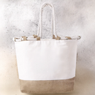 Unbedruckte Strandtasche aus natürlichen Materialien mit weißem Baumwoll-Oberteil, einem breiten Boden aus naturfarbener Jute und robusten, geflochtenen Kordelgriffen.