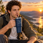 in junger Mann mit Rucksack rastet beim Wandern auf einem Berggipfel und trinkt mit einem Strohhalm aus einem großen, dunkelblauen Jwala-Thermobecher mit Henkel und dem Schriftzug „Hier und Jetzt“.